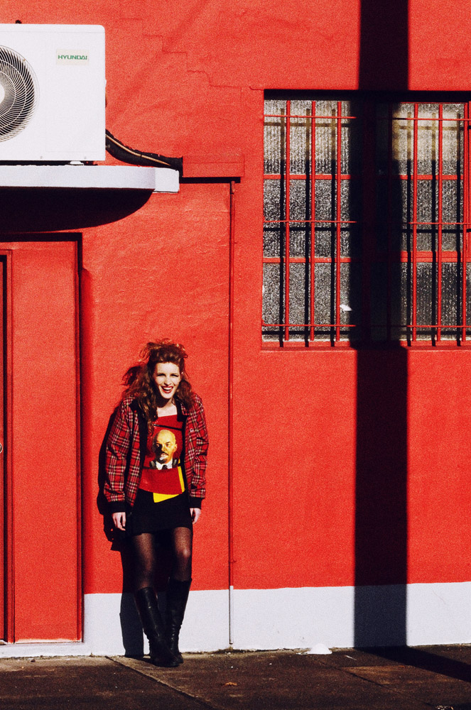 Rapture in Sydenham - Woman wearing Alicia Hollen Lenin Top & Black Ra Ra Skirt against a red wall. Tights Kmart, Boots models own. Editorial Fashion Photography by Kent Johnson.