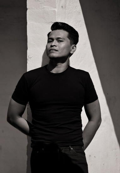 Fashion portrait of an Asian man, black tee-shirt against a white wall in a shaft of daylight.