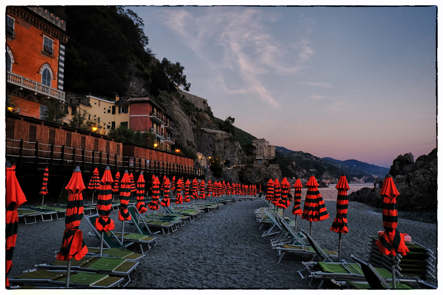 Folded orange umbrellas and green deckchairs line the beach in the evening at Monterosso al Mare, Italy. Food and travel photography by Kent Johnson