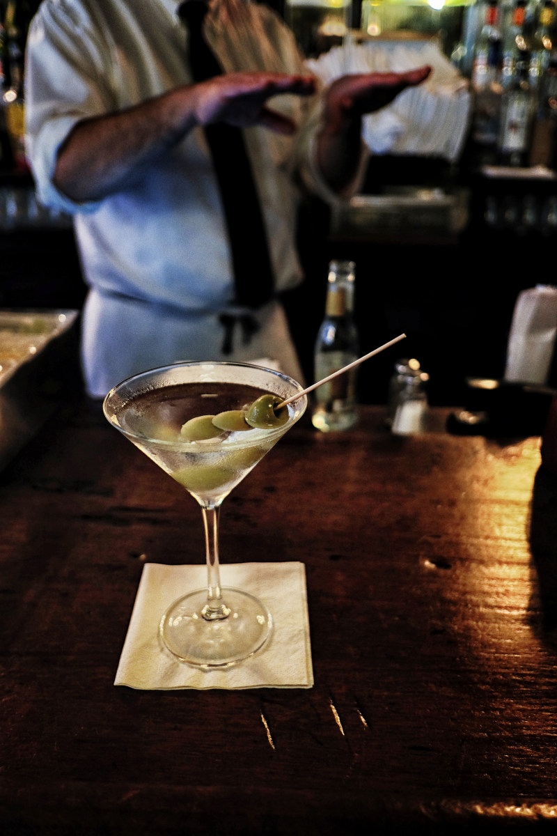 Martini with three olives on an old wooden  bar in NYC, bartenders hands behind